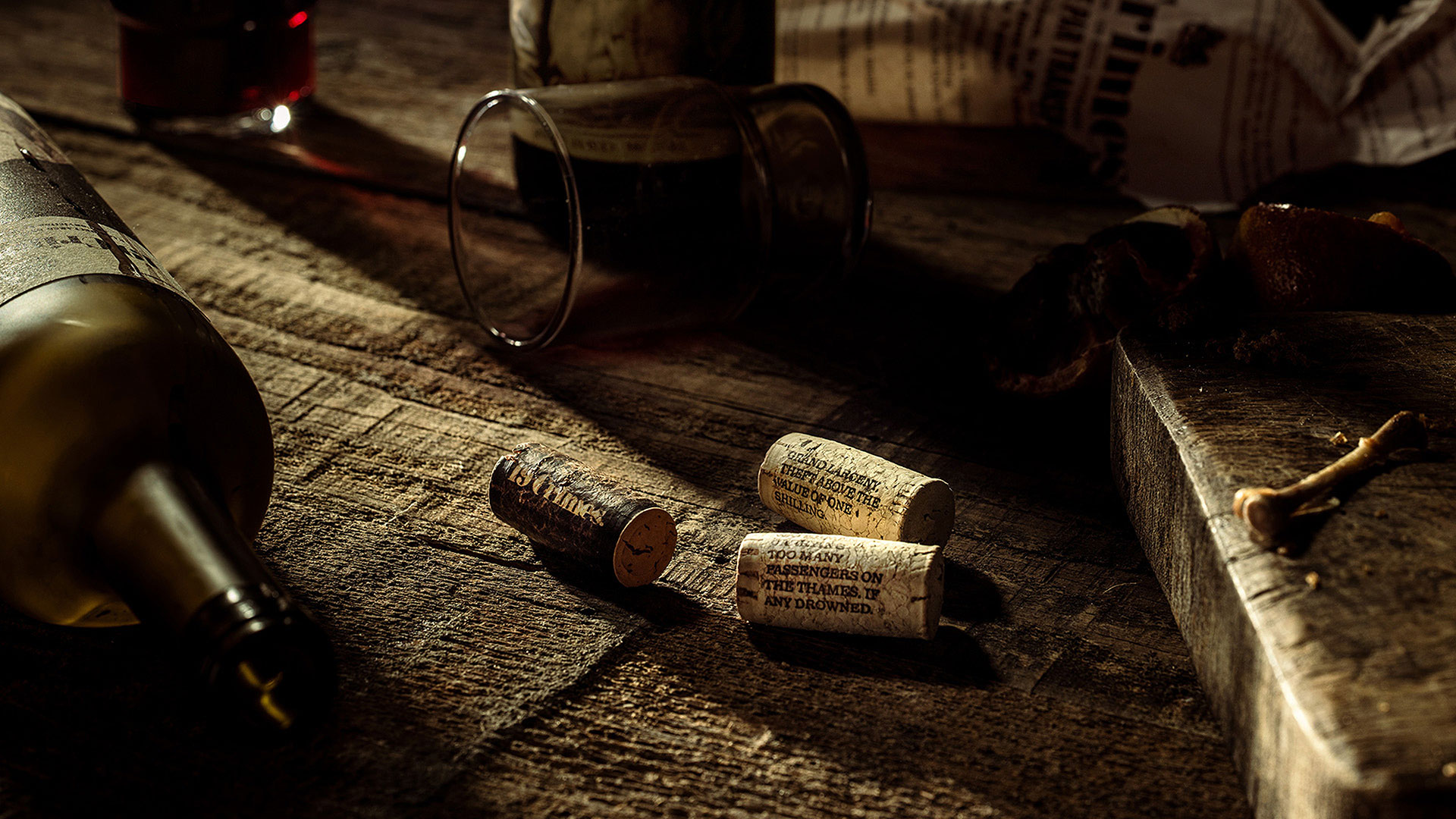 A wooden table with three wine corks, a tipped glass with red wine, a wine bottle, and assorted items in moody lighting.