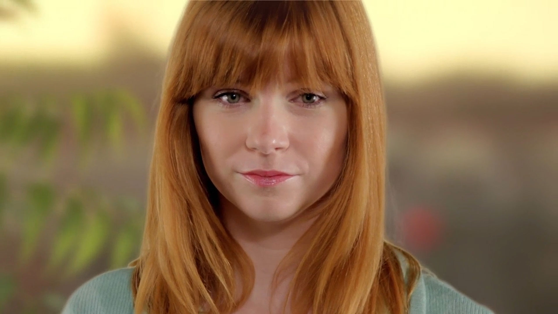 A young person with straight red hair and bangs is looking directly at the camera, with a neutral expression and a soft, blurred background.