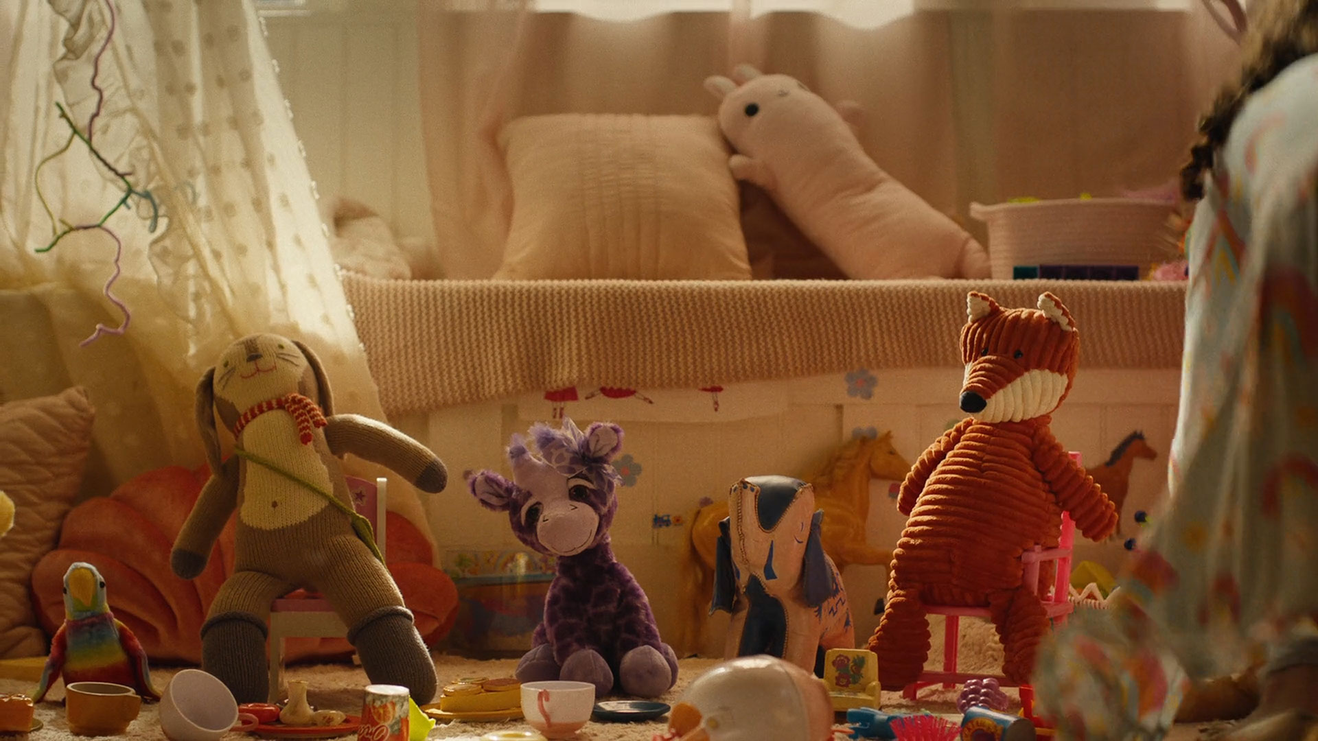 Four plush animal toys are seated in a row on small chairs in a cozy, softly lit children's playroom with pillows and various toys scattered around.
