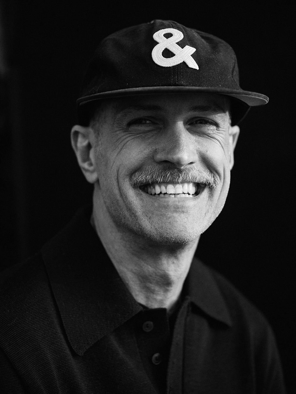 A man with a mustache smiles at the camera, wearing a black shirt and a Singlethread baseball cap with an ampersand symbol. The photo is captured in striking black and white.