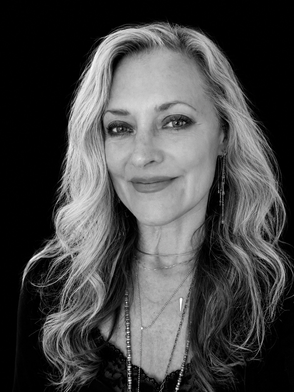 A woman with long, wavy hair smiles slightly, wearing layered necklaces and earrings—a look that evokes the understated elegance of Singlethread—set against a plain black background.