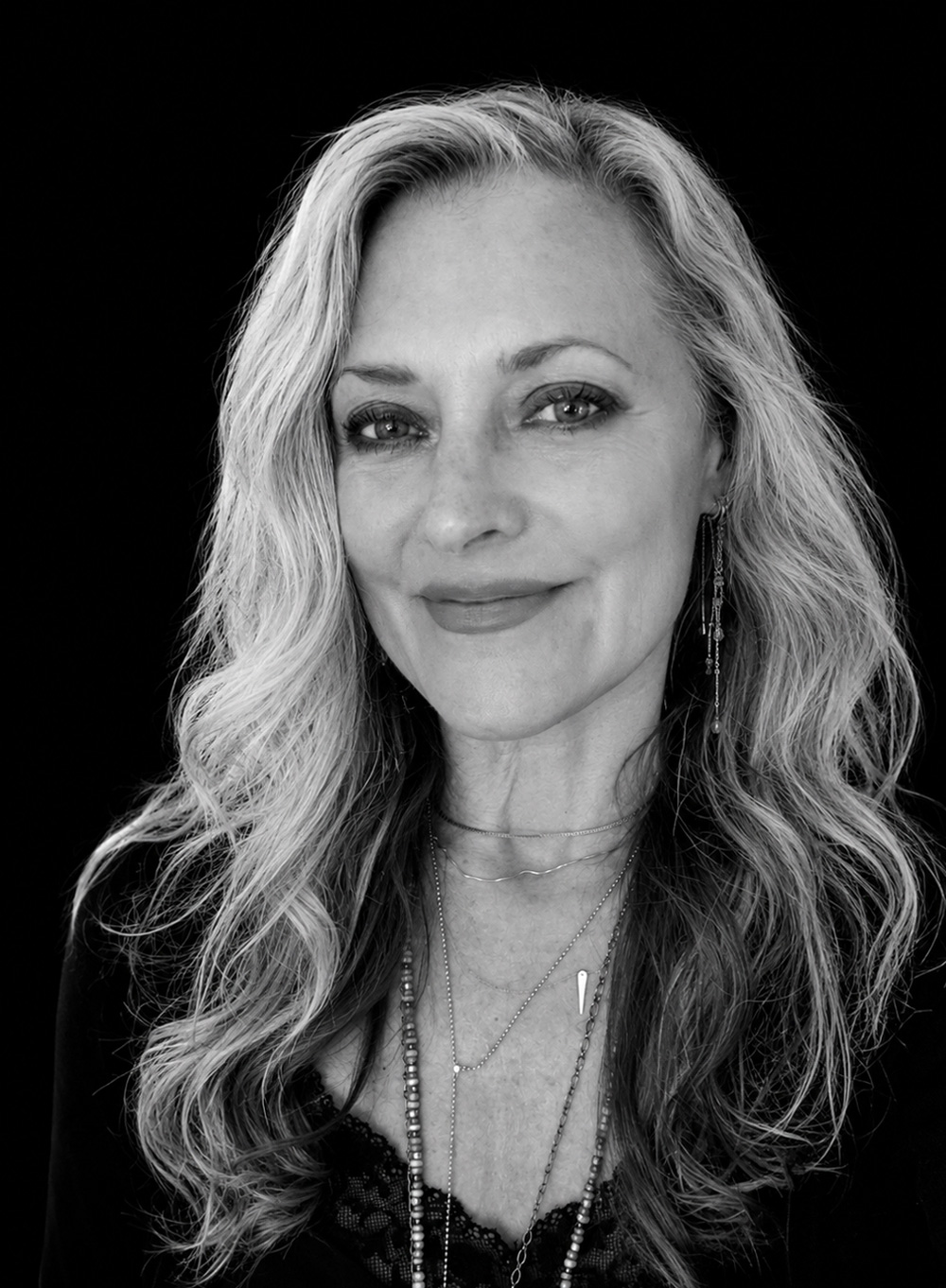 A woman with long, wavy hair smiles slightly, wearing layered necklaces and earrings&mdash;a look that evokes the understated elegance of Singlethread&mdash;set against a plain black background.