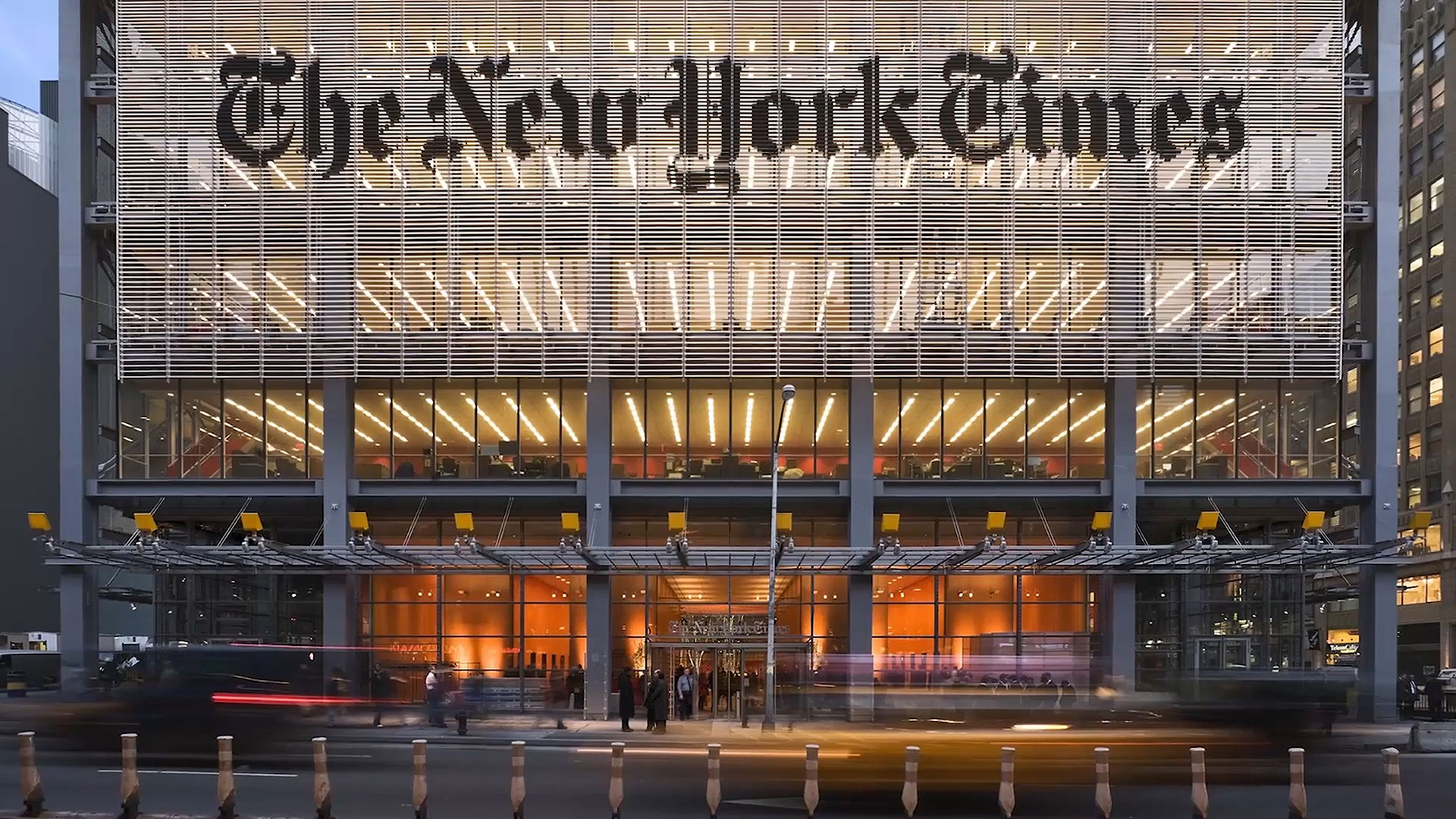 The front entrance of The New York Times building, featuring large glass doors, illuminated interior lights, and the newspaper's name displayed prominently above.