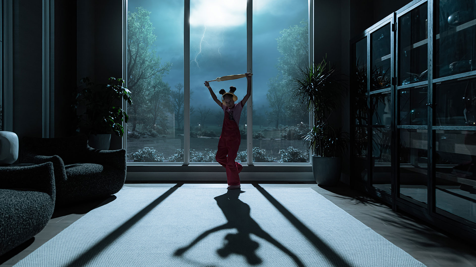 A child in pink overalls stands holding a wooden stick above their head in a living room at night, with lightning flashing through large Pella Windows behind them.