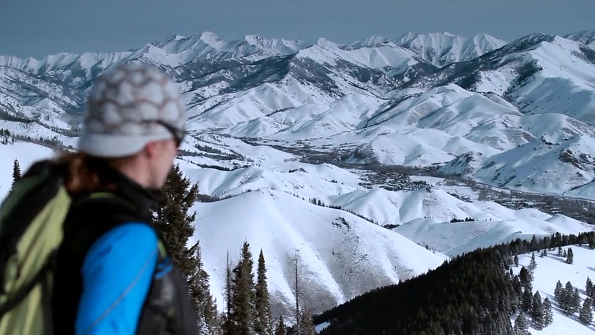 A person in winter clothing stands in the foreground, looking at a panorama of snow-covered mountain peaks and forested hills.